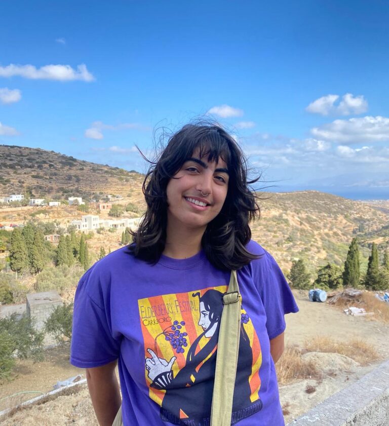 Meera Butalia smiling and standing on an overlook with a hilly town in the background.